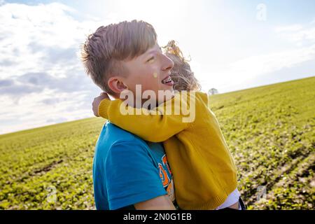 Fratello e sorella giocano all'esterno. la ragazza tiene il fratello maggiore al collo e ride felicemente. Bambini emotivi sullo sfondo di germogli di soia giovani Foto Stock