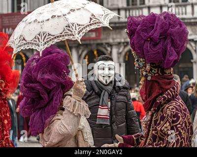 Carnevale di Venezia. Un uomo in maschera anonima è in posa con una donna mascherata in costume con un ombrellone e un uomo mascherato in costume con un cappello con gemme Foto Stock
