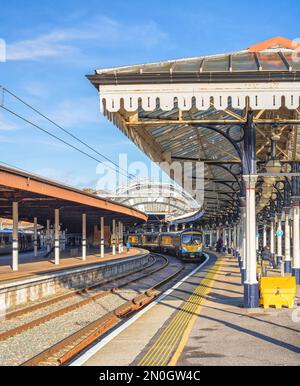 Vista di una stazione ferroviaria con rotaie che conducono verso un arco di ferro. Una piattaforma è coperta da una tettoia del 19th ° secolo e un treno si trova accanto a un plateau Foto Stock