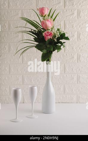 Composizione STILL LIFE composta da bottiglia di champagne bianco, bicchieri e bouquet di fiori rosa sul tavolo. Sfondo bianco festa. San Valentino Foto Stock