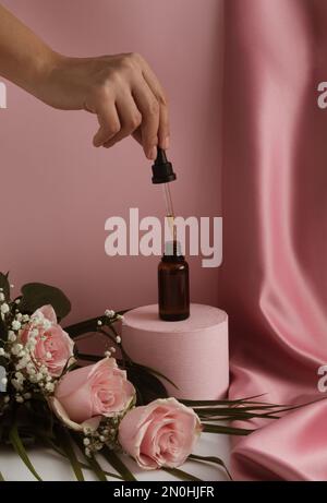 Bottiglia contagocce in vetro marrone su un podio, fiori rosa e mano della donna. Sfondo tenda di seta rosa Patel. Prodotti per la cura della pelle, cosmetici naturali. B Foto Stock