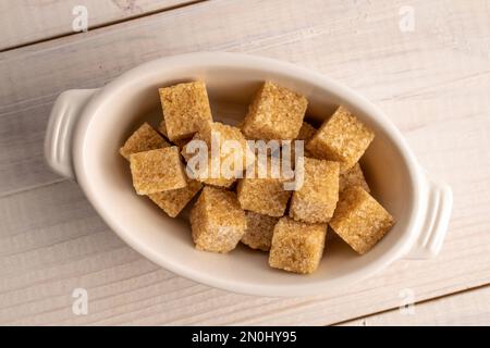 Diversi cubetti di zucchero di canna in una ciotola di ceramica bianca, primo piano, su un tavolo di legno bianco. Foto Stock