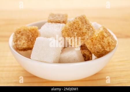 Diversi cubetti di zucchero di canna e zucchero bianco in una ciotola bianca in ceramica, primo piano, su un tavolo di legno. Foto Stock