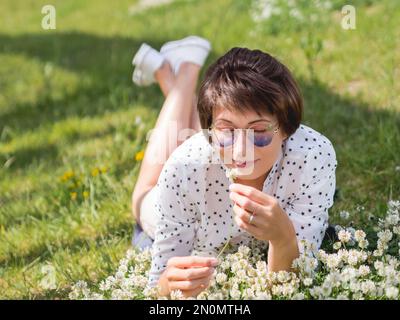 Donna in occhiali da sole colorati sniffs fiori trifoglio sul prato in parco urbano. Natura in città. Rilassati all'aperto dopo il lavoro. Atmosfera estiva. Foto Stock