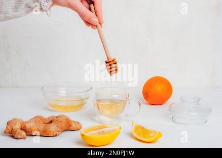 Bicchiere con tè all'arancia, al limone e allo zenzero e miele che si versa in una tazza. Ingredienti su tavola di legno bianco. Foto Stock