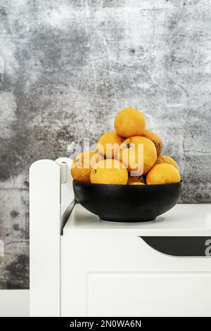 Ciotola di frutta nera piena di mandarini maturi su una panca bianca Foto Stock