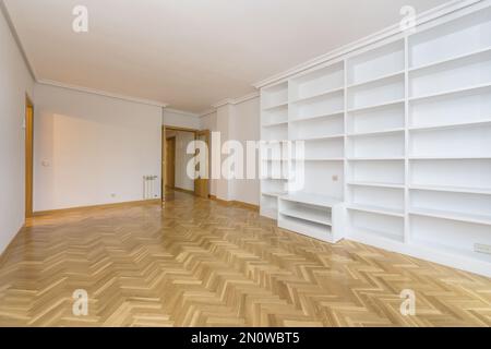Camera vuota con una parete coperta da una libreria in muratura dipinta di bianco, pavimenti in parquet di quercia francese laccato e lucidato disposti a spina di pesce a Foto Stock