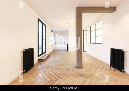 Camera vuota di un appartamento in stile loft con pareti dipinte di bianco, radiatori in ghisa nera, finestre in ferro con divisori in vetro e una colonna di cemento grezzo Foto Stock