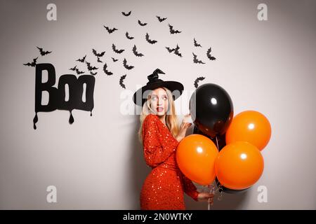 Donna in cappello di strega con i palloncini che posano vicino alla parete bianca decorata per Halloween Foto Stock