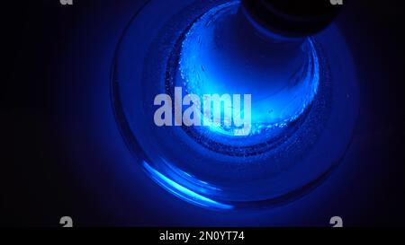 Primo piano di una borraccia trasparente di gancetta di vetro sul tavolo con una nuvola di fumo. Borraccia di narghilè sotto la lampada blu che si erge su un tavolo di legno Foto Stock