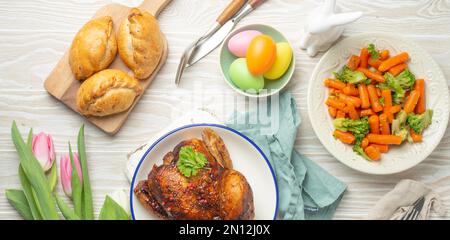 Cibo pasquale su bianco tavolo rustico: Uova color pastello, pollo arrosto e verdure, panini e fiori primaverili tulipani vista dall'alto fieno lay, Pasqua famil Foto Stock