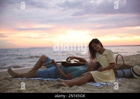 Coppia deliziosa con chitarra e cestino da picnic sulla spiaggia al tramonto Foto Stock