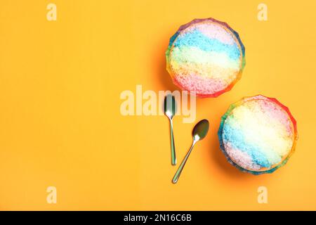 Rasatore di ghiaccio arcobaleno in ciotole di vetro da dessert e cucchiai su fondo arancione, piatto. Spazio per il testo Foto Stock