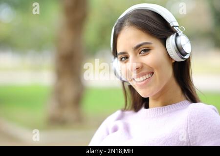 Una donna felice che indossa le cuffie guarda la macchina fotografica seduta in un parco Foto Stock