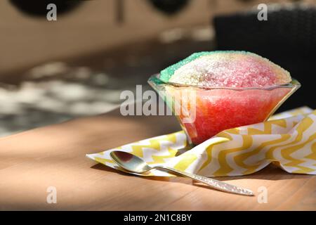 Rasatore di ghiaccio arcobaleno in ciotola di vetro da dessert e cucchiaio su tavolo di legno all'aperto Foto Stock