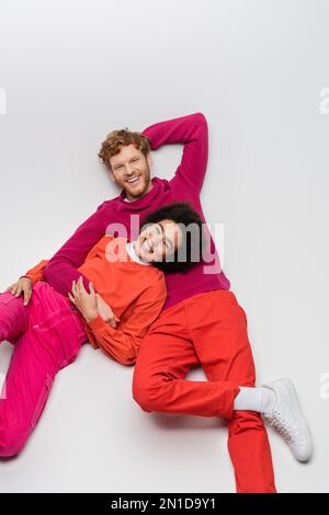 vista dall'alto di una allegra donna afro-americana che riposa con un uomo rosso in abiti color magenta su bianco, immagine stock Foto Stock