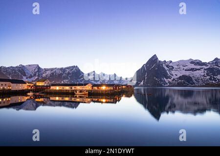 Villaggio di pescatori che si riflette nel mare al crepuscolo con montagne innevate sullo sfondo, Hamnoy, Reine Bay, Isole Lofoten, Norvegia, Scandinavia, Europa Foto Stock