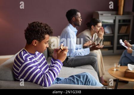 Ragazzo adolescente che gioca su smartphone quando i suoi genitori parlano con psicologo in background Foto Stock