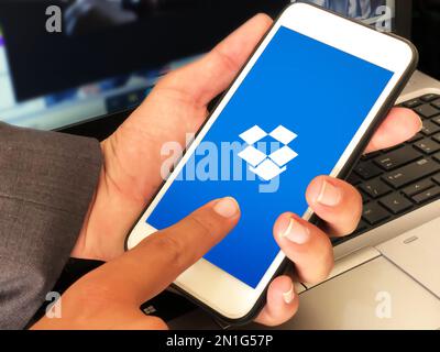 Uomo che utilizza dropbox di applicazioni di archiviazione su dispositivi mobili con laptop sullo sfondo, background tecnico editoriale Foto Stock
