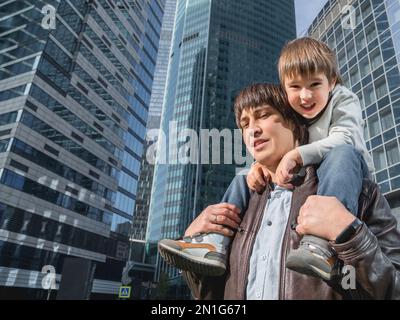 Il ragazzino si siede sulle spalle del padre tra i grattacieli. Papà e figlio guarda su pareti di vetro di edifici. Tecnologie future e moderne, equilibrio di vita Foto Stock