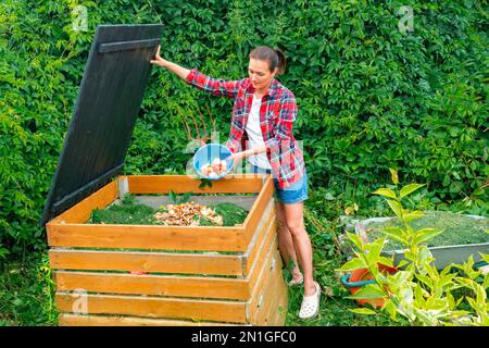 Spreco di cibo in un bidone di compost. Una donna in una maglietta a quadri getta rifiuti da cucina in un bidone per compost fai da te. Invecchiamento di composto per la sua introduzione nel suolo Foto Stock