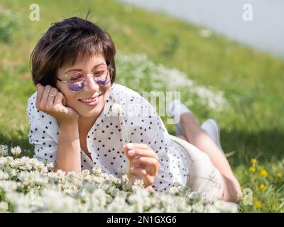 Donna in occhiali da sole colorati sniffs fiori trifoglio sul prato in parco urbano. Natura in città. Rilassati all'aperto dopo il lavoro. Atmosfera estiva. Foto Stock