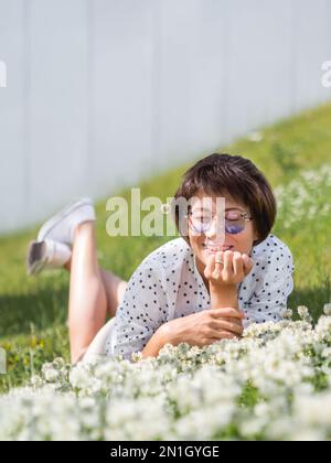 Donna in occhiali da sole colorati sniffs fiori trifoglio sul prato in parco urbano. Natura in città. Rilassati all'aperto dopo il lavoro. Atmosfera estiva. Foto Stock