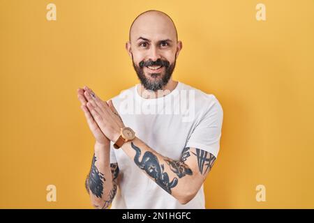 Giovane uomo ispanico con tatuaggi in piedi su sfondo giallo che batte e applaude felice e gioioso, sorridendo mani orgogliose insieme Foto Stock