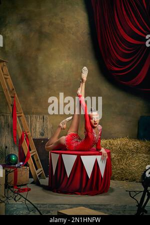 Spettacolo da circo retrò. Look grazioso. Piccola ragazza flessibile, ginnastica o acrobat in costume rosso facendo ginnastica trucco sul backstage circo scuro Foto Stock