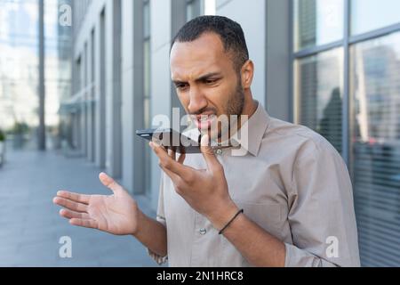 Arrabbiato uomo d'affari in camicia registrazione di messaggi audio da fuori ufficio edificio, uomo afroamericano utilizzando assistente dando comandi di intelligenza artificiale utilizzando smartphone. Foto Stock