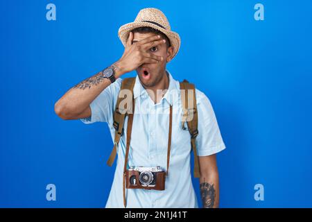 Giovane brasiliano che tiene la macchina fotografica vintage peeking in shock coprendo il viso e gli occhi con la mano, guardando attraverso le dita con l'espressione imbarazzata. Foto Stock