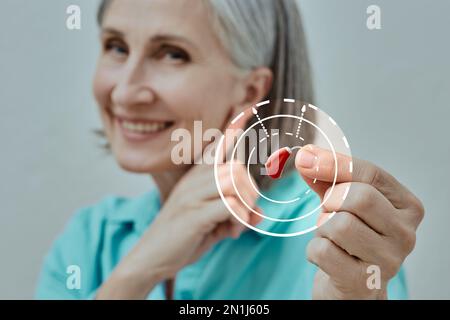 Donna matura che tiene in mano l'apparecchio acustico BTE in primo piano e punta all'orecchio, concetto. Trattamento per la perdita dell'udito Foto Stock