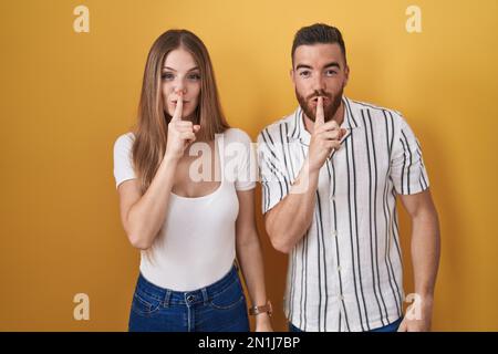 Giovane coppia in piedi su sfondo giallo chiedendo di essere tranquilla con il dito sulle labbra. silenzio e concetto segreto. Foto Stock