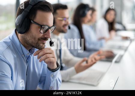 Primo piano di un bell'addetto dell'assistenza clienti maschile con cuffie che lavorano nel call center. Gruppo di operatori helpdesk al servizio di assistenza clienti Foto Stock