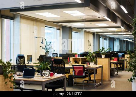 Parte di uno spazioso e contemporaneo ufficio nel centro affari con lungo corridoio lungo l'area di coworking con fila di scrivanie con sedie e piante verdi Foto Stock