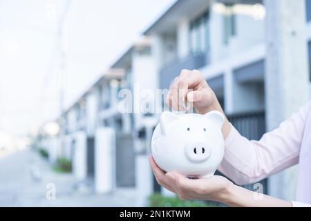 Risparmio di denaro per acquistare casa, mani risparmio di denaro in piggy banca di fronte al bene immobile. Foto Stock
