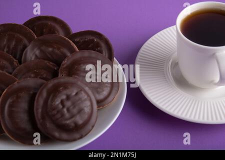 Foto dei biscotti immersi nel cioccolato, sdraiati su un piatto con una tazza di caffè accanto su uno sfondo viola Foto Stock