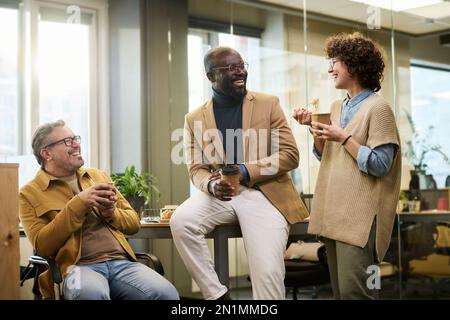 Gruppo di allegri gestori di uffici interculturali che hanno caffè e wok cinese e ridendo di cose divertenti mentre si gode la pausa pranzo Foto Stock