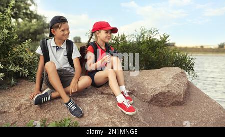 Carini bambini con zaini seduti sulla roccia vicino al fiume. Viaggio in campeggio Foto Stock