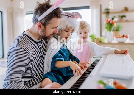 I bambini felici in costumi pasquali suonano al pianoforte, con il loro papà. Foto Stock