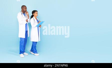 Pensivo afro-americano uomo e donna medici in camici bianchi, pensando, guardando lo spazio libero, sfondo blu panorama Foto Stock