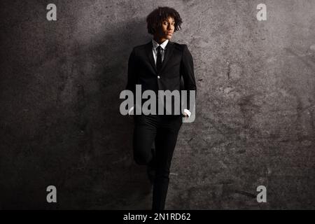 Giovane modello afro-americano maschile in tuta e cravatta appoggiato su un muro arrugginito scuro Foto Stock