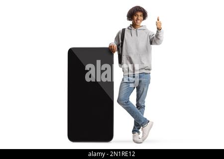 Giovane studente afro-americano appoggiato su un grande telefono cellulare e gesturing pollici isolato su sfondo bianco Foto Stock