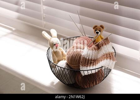 Palle di filato, aghi per maglieria e graziosi giocattoli fatti a mano in un cestino di metallo sul davanzale interno. Hobby creativo Foto Stock