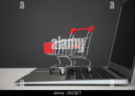 Shopping su Internet. Computer portatile con carrello piccolo sul tavolo su sfondo grigio Foto Stock