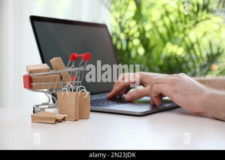 Shopping su Internet. Piccolo carrello con scatole e borse vicino a un uomo che utilizza un computer portatile al chiuso Foto Stock