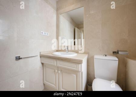 Bagno con ripiano in marmo color crema su mobili in legno bianco, specchio integrato nella parete e piastrelle dello stesso materiale Foto Stock