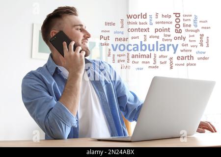 Giovane uomo che parla al telefono cellulare mentre lavora con un computer portatile alla scrivania, parola nuvola davanti a lui Foto Stock