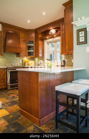 Cucina con pavimento in ardesia dai toni terrosi, armadi in legno di ciliegio e isola con ripiano in quarzo bianco all'interno di una casa in stile cottage contemporaneo. Foto Stock