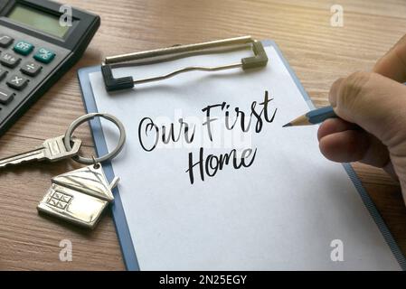 Messa a fuoco selettiva della calcolatrice, chiave della casa, una matita che tiene la mano che scrive una frase " la nostra prima casa" su una clipboard con carta su sfondo di legno. Foto Stock